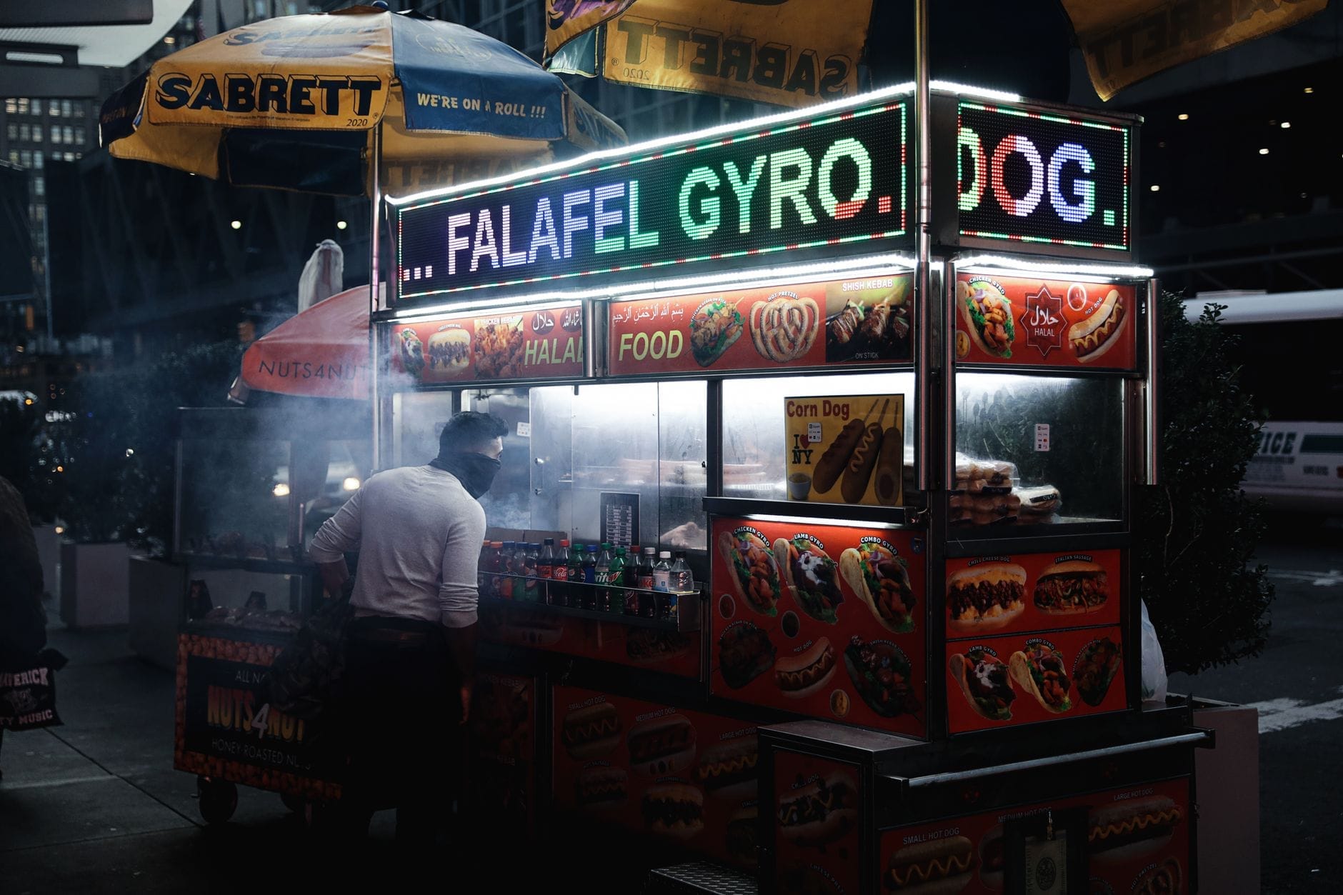 Carrinho de comida de rua clássico de Nova York à noite, com letreiros iluminados de falafel e gyro, rodeado de vapor e luzes da cidade.