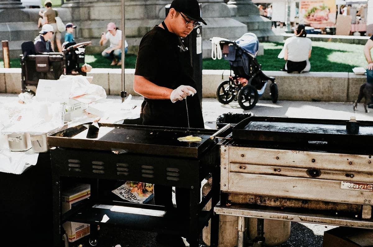 Outdoor-Essensverkäufer beim Grillen auf einem belebten Markt mit Menschen, Kinderwagen und Grünpflanzen im Hintergrund an einem sonnigen Tag.