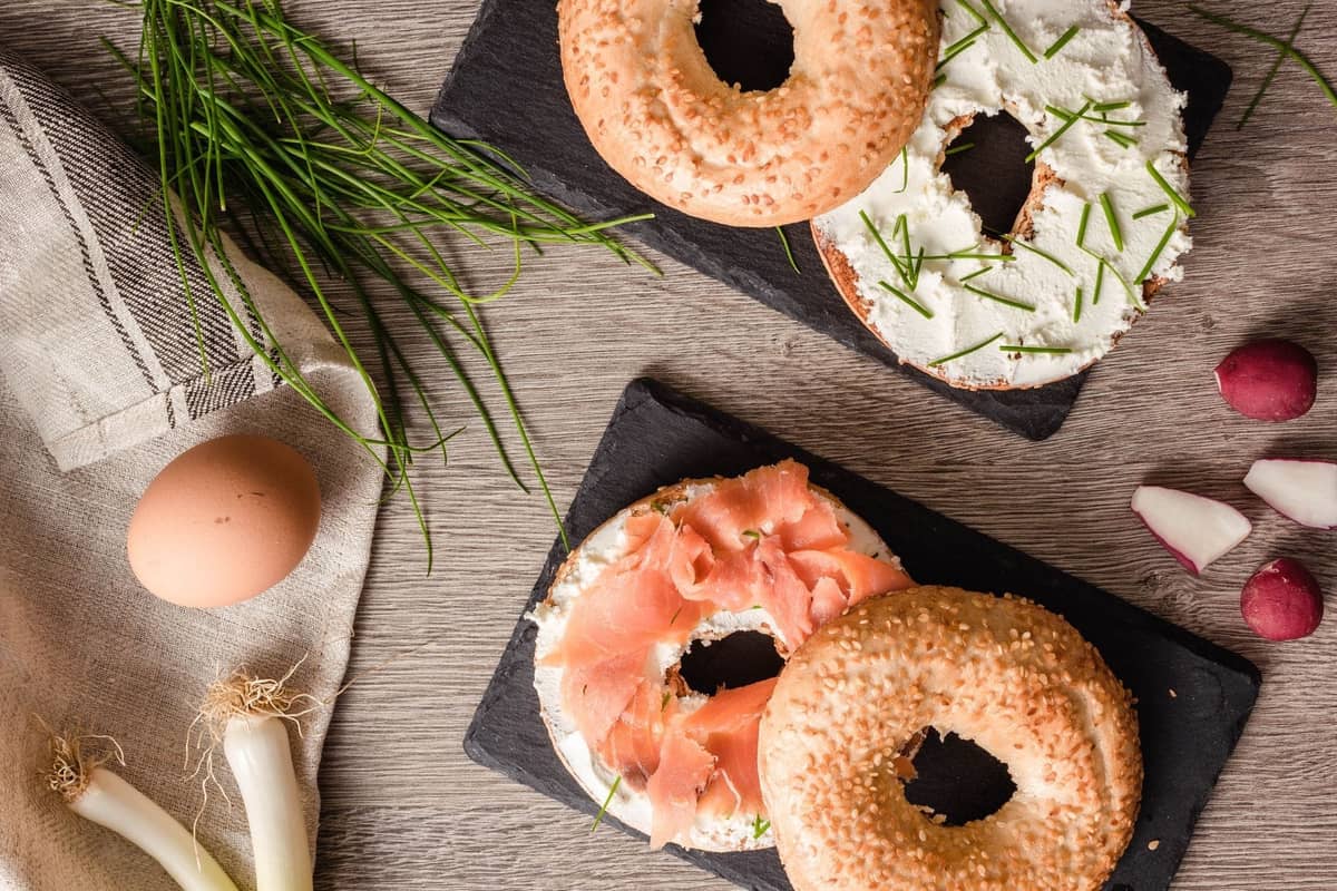 Zwei Bagels, einer mit Lox, Frischkäse und Schnittlauch, auf schwarzen Schieferplatten, umgeben von frischen Zutaten
