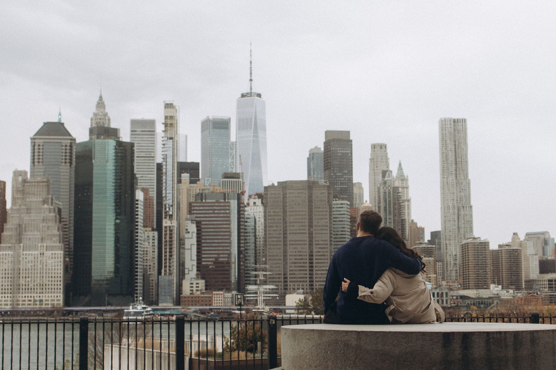 Um casal se abraça em um banco com vista para o icônico skyline de Manhattan, com arranha-céus imponentes e a orla visíveis em um dia nublado.