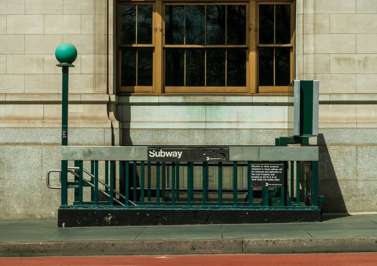 ทางเข้าสถานีรถไฟใต้ดินคลาสสิกของ New York City พร้อมโคมไฟทรงกลมสีเขียว ราวบันได และป้ายรถไฟใต้ดินหน้าอาคารหิน