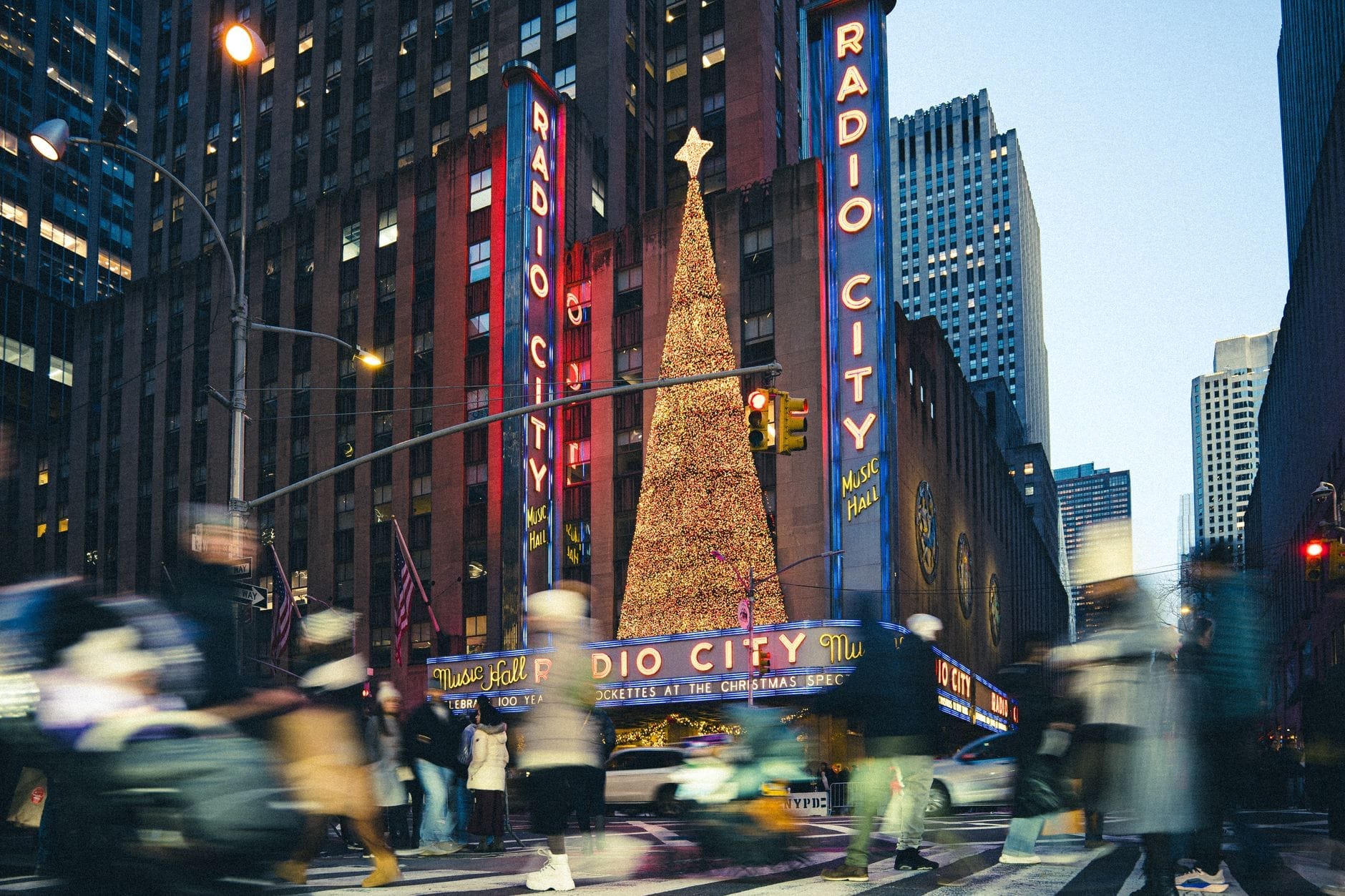 Multidão atravessa uma movimentada rua do Midtown com o Radio City Music Hall e uma enorme árvore de Natal iluminada ao fundo.