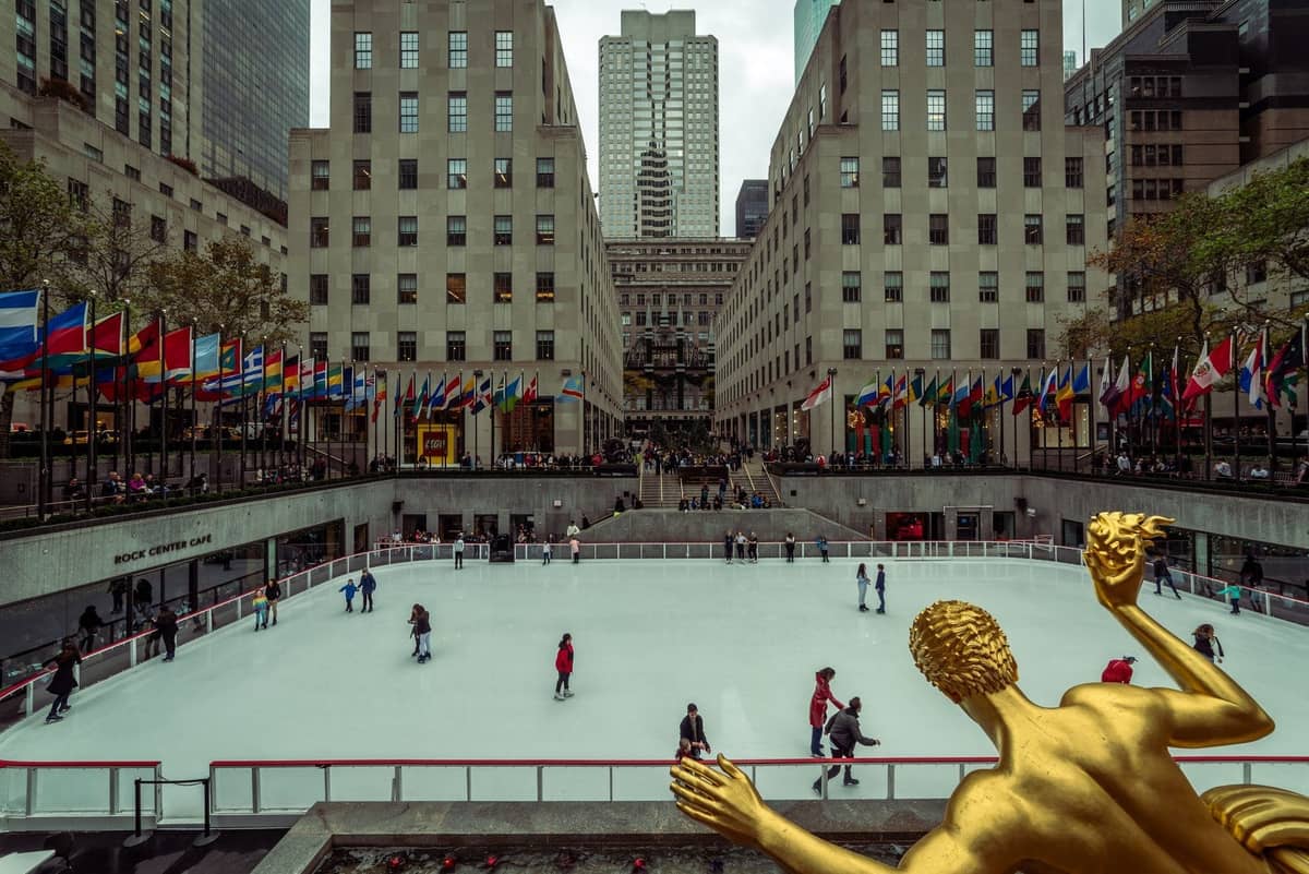 Tagesansicht der Eislaufbahn des Rockefeller Center mit Schlittschuhläufern, goldener Prometheus-Statue, Flaggen und umliegenden Gebäuden in New York City.