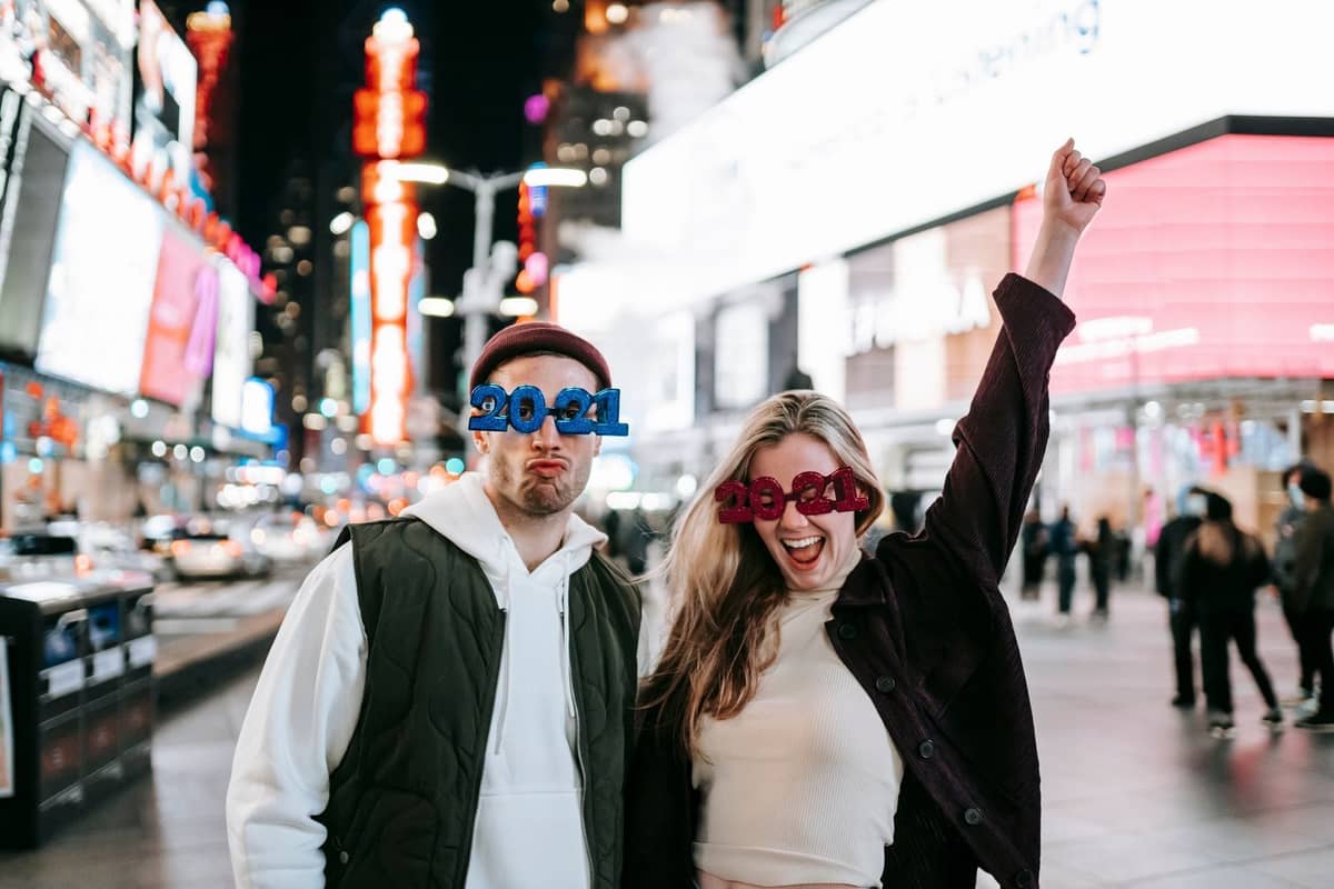 Zwei Personen mit 2021-Brillen feiern nachts am Times Square, umgeben von hellen Stadtlichtern und Leuchtreklamen.