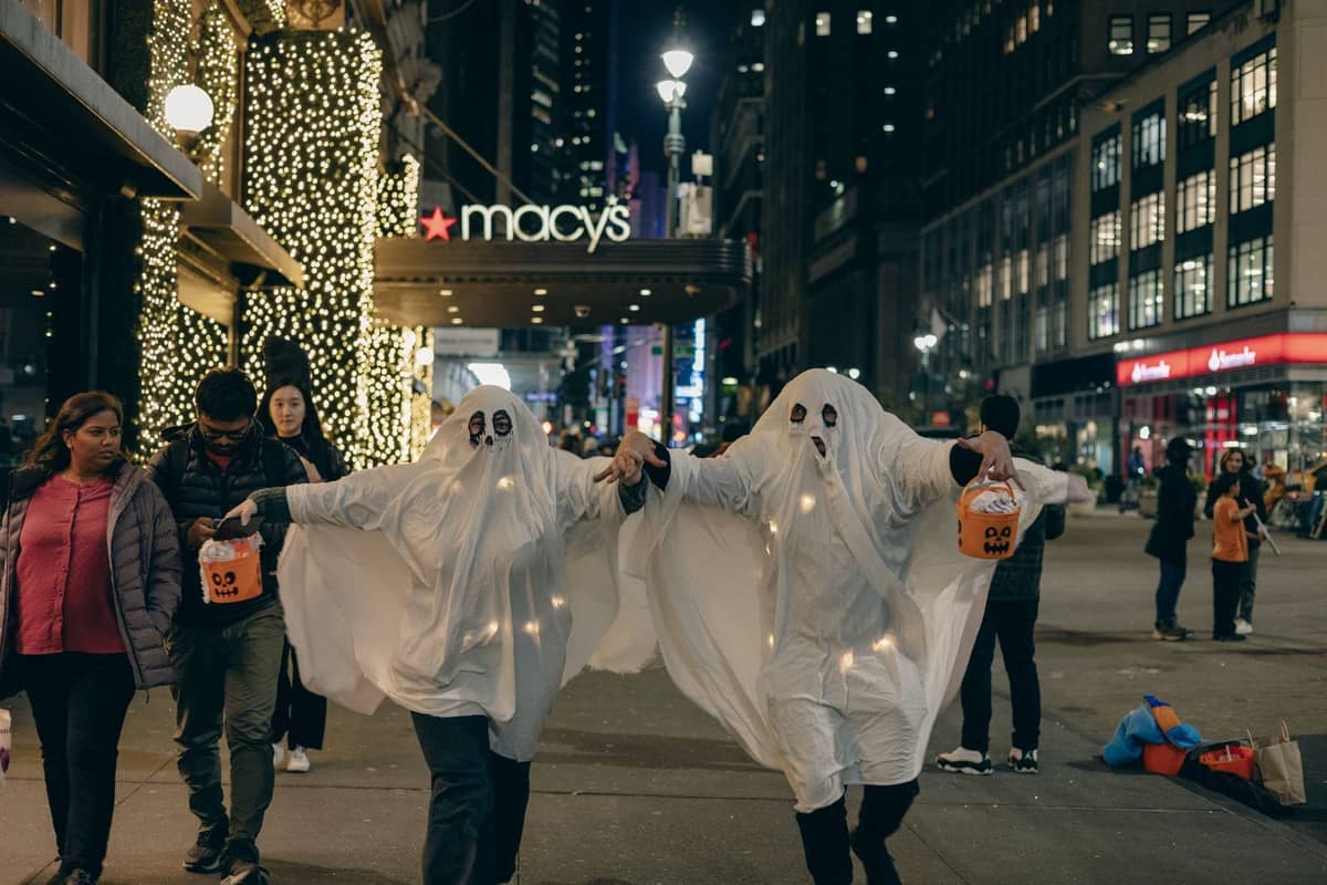 Menschen in Geisterkostümen beim Trick-or-Treating in der Nähe von Macy's in New York City bei Nacht, mit festlichen Lichtern und Stadtmenschenmassen.