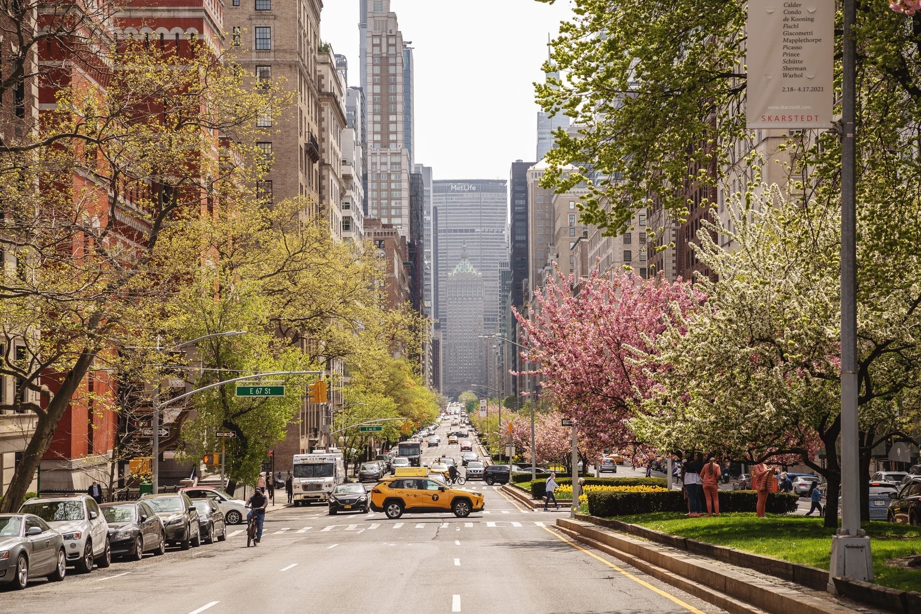 Uma rua ensolarada de Nova York na primavera com árvores floridas, o clássico táxi amarelo, pessoas caminhando e arranha-céus ao fundo.