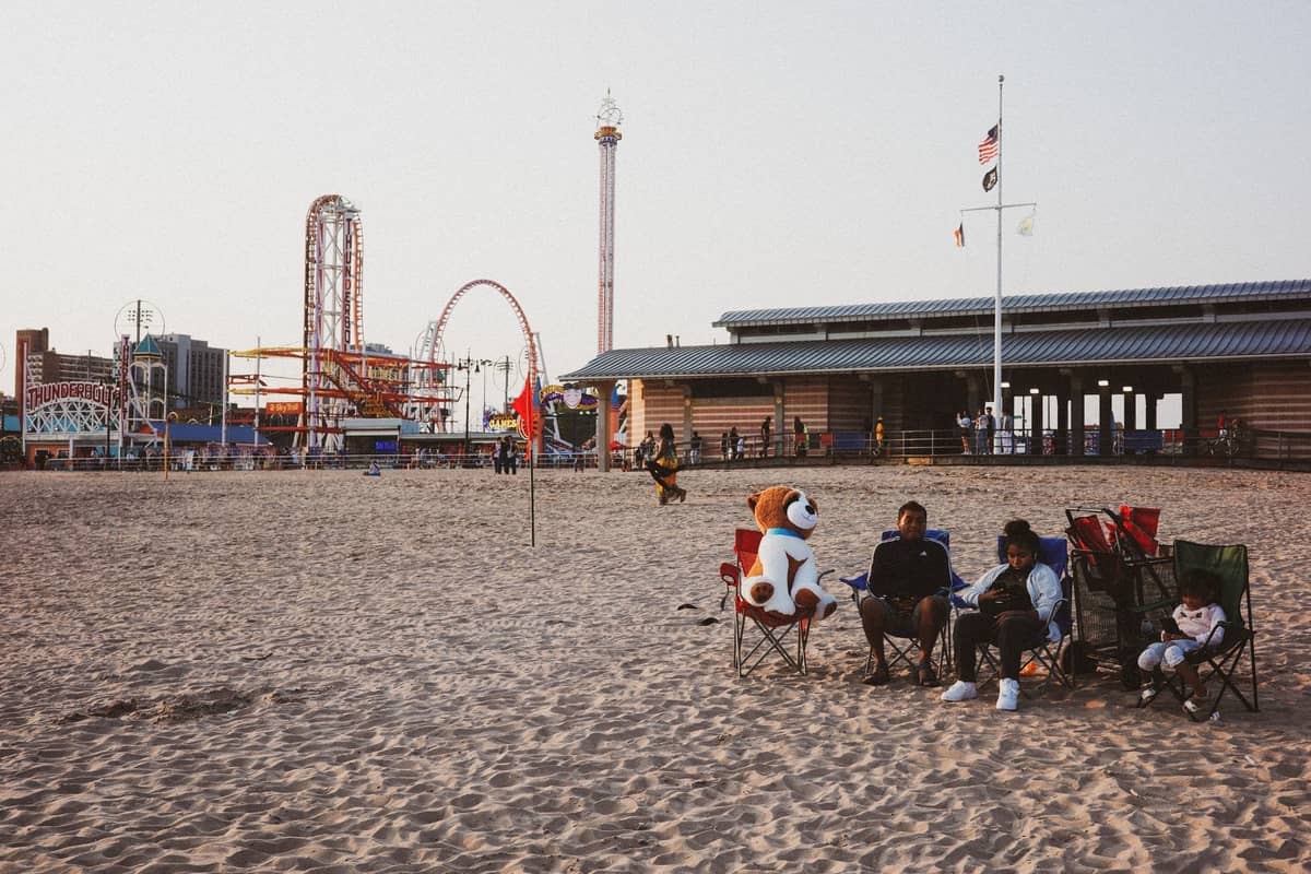 Menschen entspannen sich am sandigen Strand von Coney Island mit Fahrgeschäften eines Vergnügungsparks und Boardwalk-Gebäuden im Hintergrund, New York City.