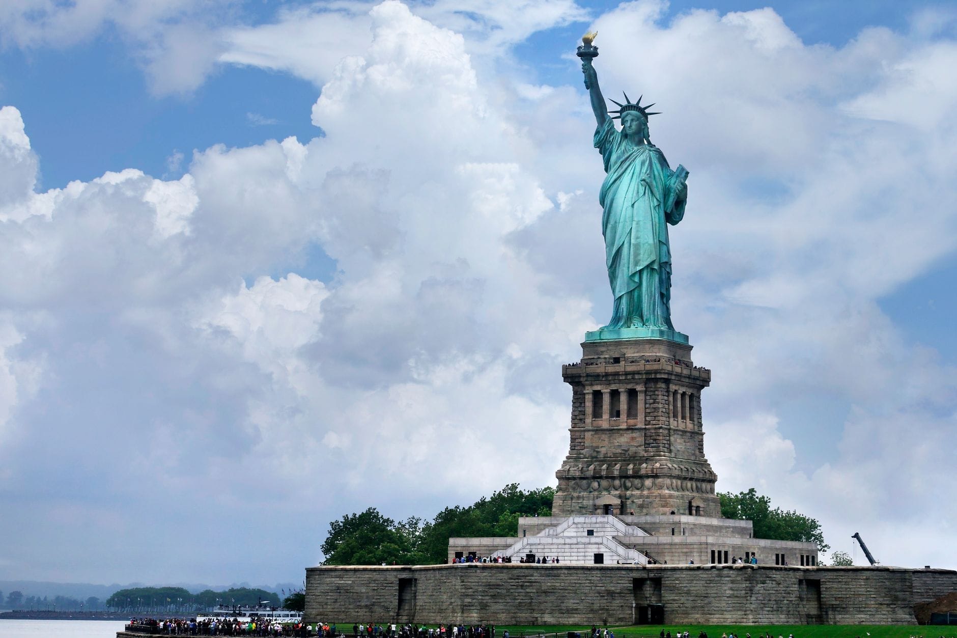 A Estátua da Liberdade se ergue imponente sob um céu parcialmente nublado de verão, com multidões de visitantes e vegetação exuberante ao seu redor em Nova York.