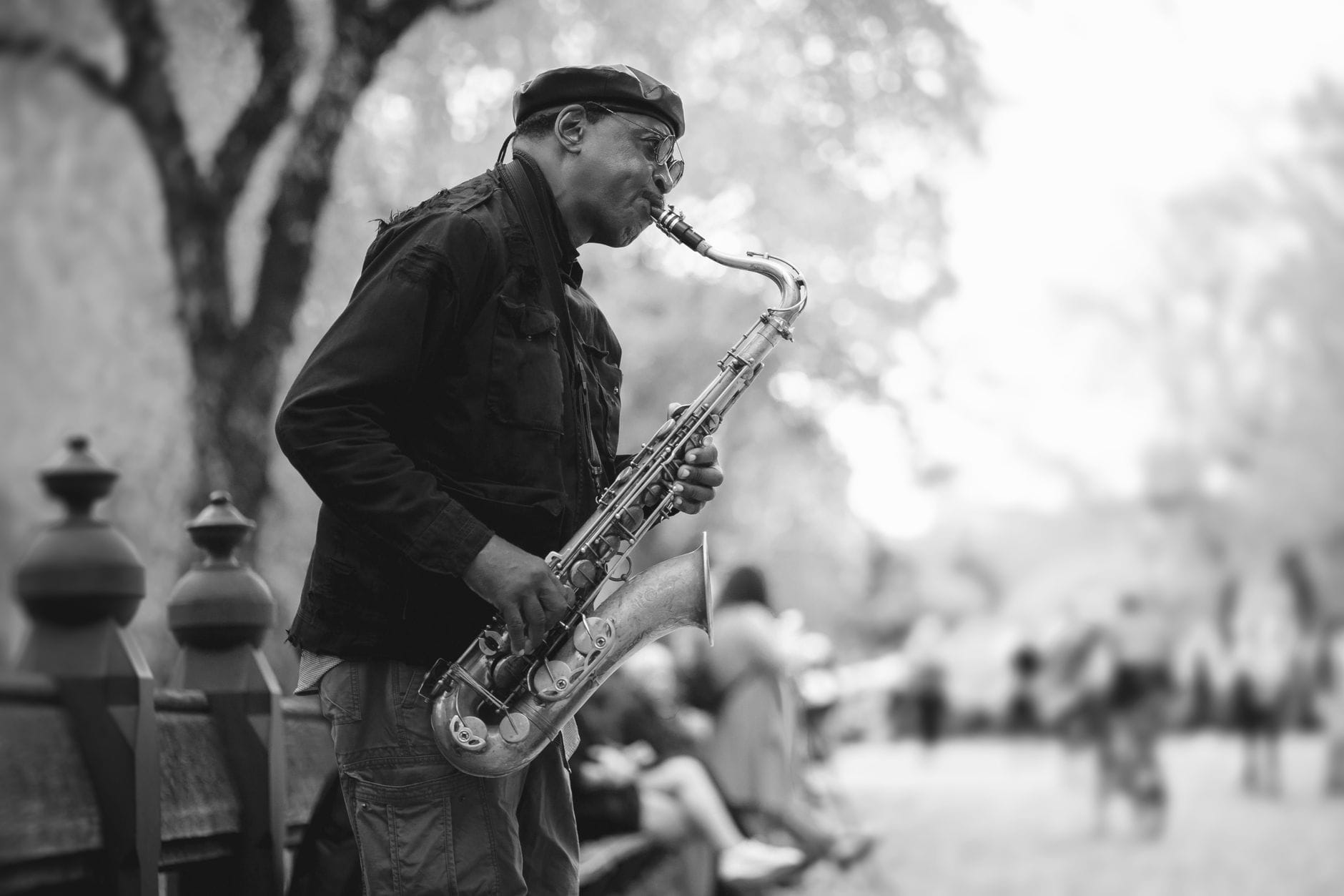 Foto em preto e branco de um saxofonista de jazz tocando ao ar livre numa rua de Nova York, com pessoas e árvores desfocadas ao fundo.