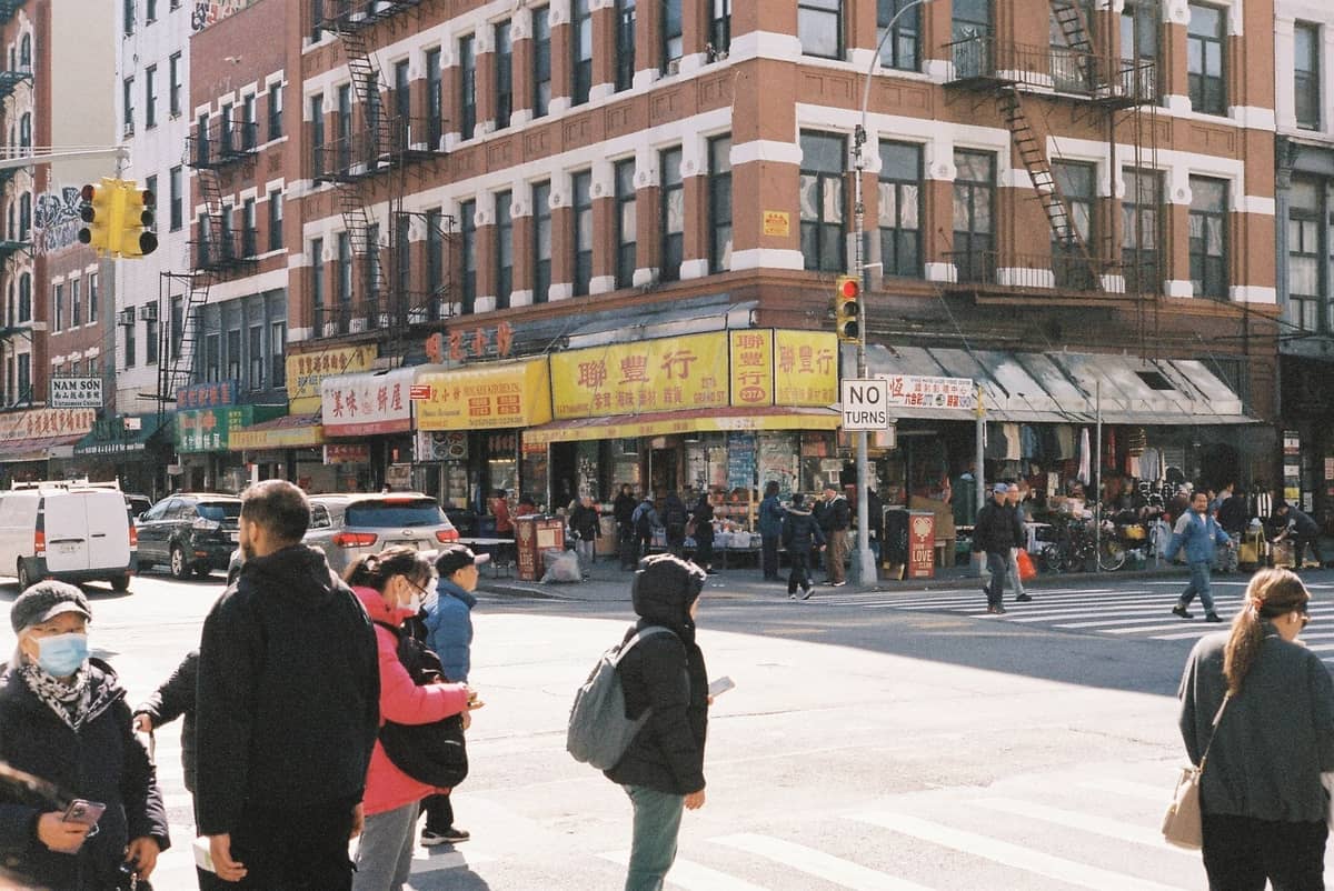 Belebte Straßenecke in New York City mit überquerenden Fußgängern, asiatischen Geschäftsfronten und Schildern in einem vielfältigen Stadtviertel.