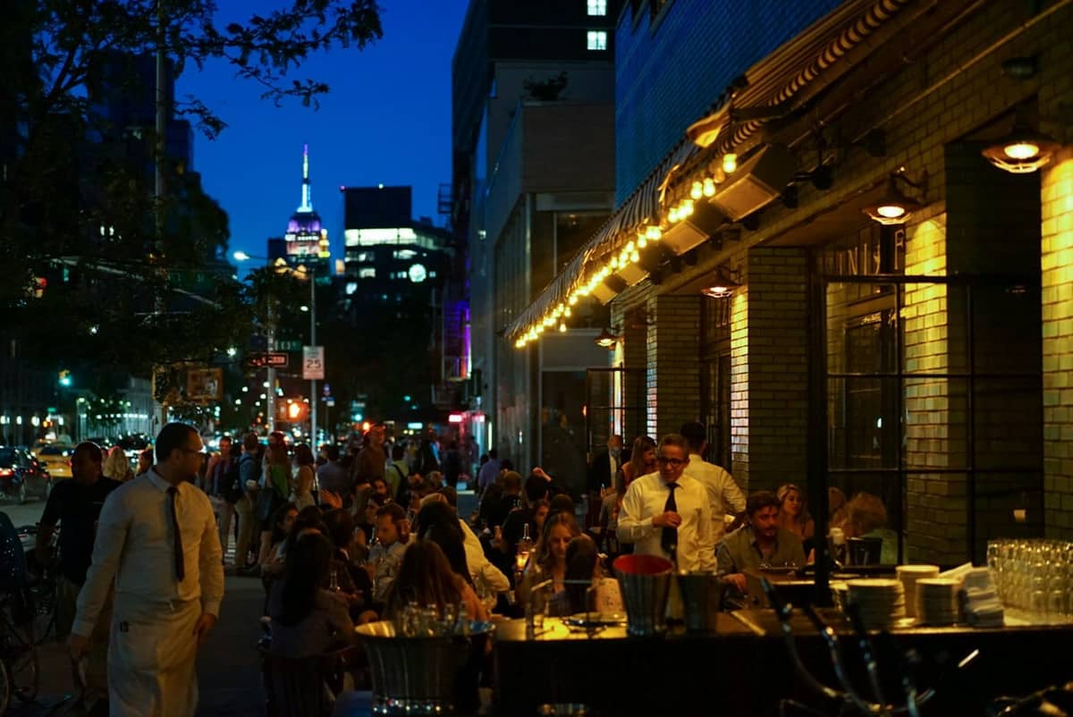 โต๊ะนั่งกลางแจ้งที่บาร์คึกคักใน New York City ยามค่ำคืน พร้อมผู้คนรับประทานอาหารและแสงไฟของเมืองในฉากหลัง