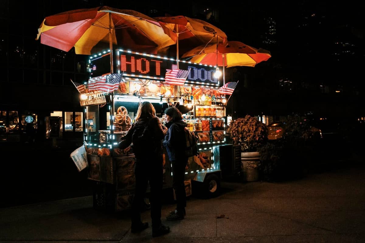 สองคนกำลังสั่งอาหารจากรถขายฮอตด็อกที่สว่างไสวในยามค่ำคืนบนทางเท้าของ New York City