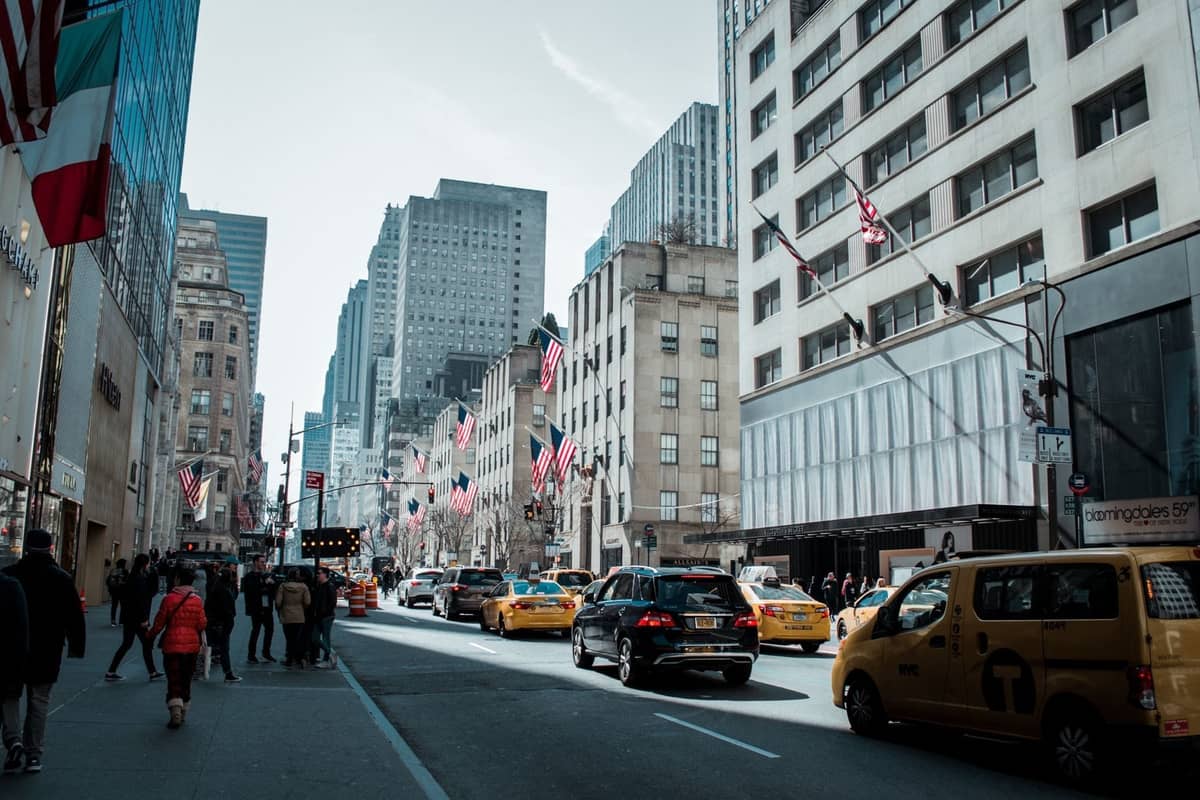 ถนน Fifth Avenue ใน Midtown Manhattan พร้อมธงชาติอเมริกัน นักช้อปปิ้ง แท็กซี่สีเหลือง และร้านค้าหรูหราที่เรียงรายอยู่ตามถนนที่คึกคัก