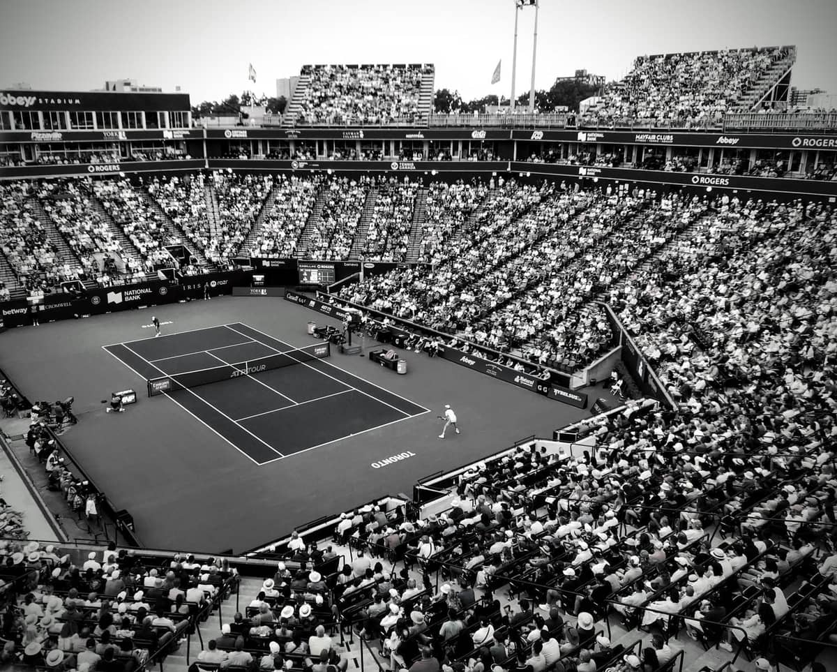 Schwarz-Weiß-Bild eines großen, überfüllten Tennisstadions von oben während eines Spiels, mit vollen Rängen und Spielern auf dem Platz.
