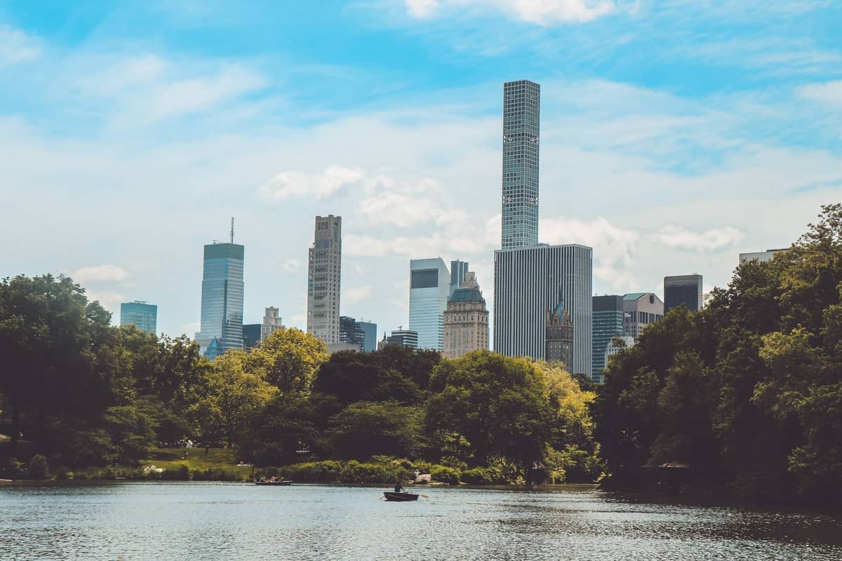 Blick auf den See im Central Park mit üppig grünen Bäumen und der Skyline von New York City im Hintergrund an einem sonnigen Tag.
