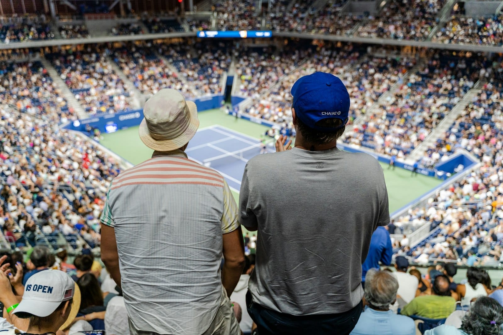Estádio de tênis lotado durante o US Open, com dois espectadores em primeiro plano, quadra visível e a atmosfera vibrante do torneio.