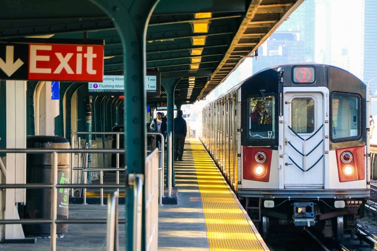 Ein New York City 7-Zug, der an einem erhöhten Bahnsteig ankommt, mit wartenden Fahrgästen und sichtbaren Ausgangsschildern.