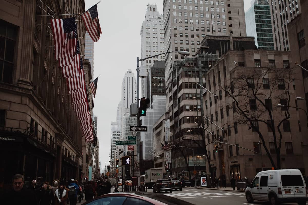 วิวของ Fifth Avenue ใน Midtown Manhattan พร้อมธงชาติอเมริกา ถนนที่คึกคัก ตึกระฟ้า และผู้คนเดินเท้าในวันที่มีเมฆมาก