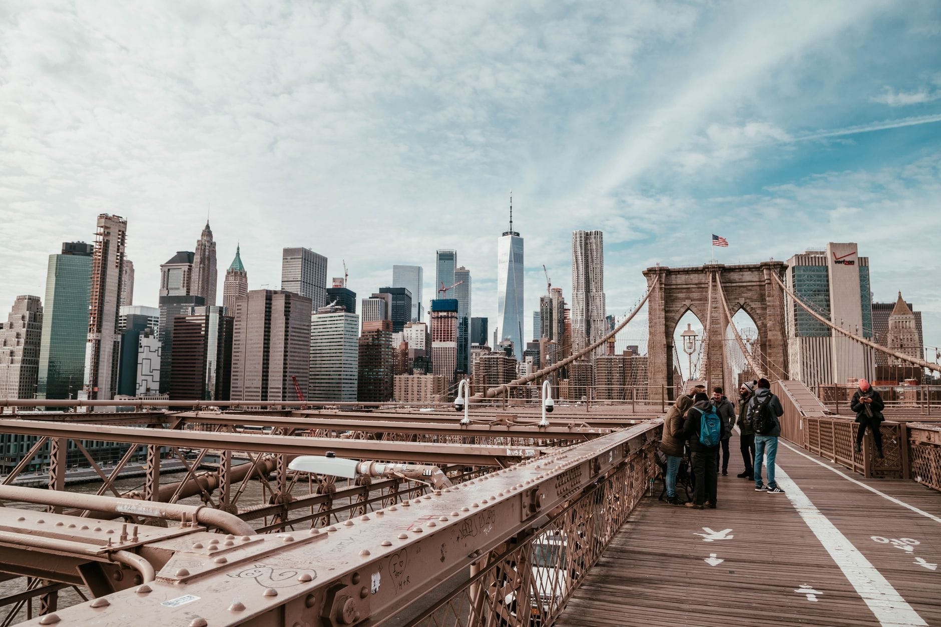 Pessoas caminhando pela Ponte do Brooklyn em Nova York com o skyline de Manhattan ao fundo em um dia parcialmente nublado.