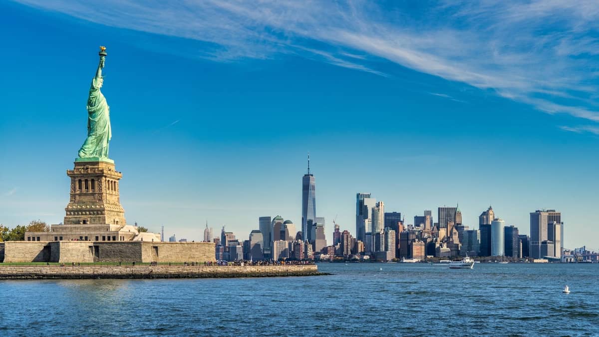 อนุสาวรีย์เทพีเสรีภาพในเบื้องหน้า พร้อมเส้นขอบฟ้าของ Lower Manhattan และ One World Trade Center ในวันที่แจ่มใสและแดดจัด