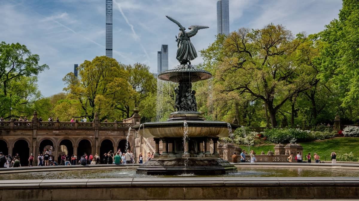 วิวกว้างของ Bethesda Fountain และ Terrace ใน Central Park พร้อมผู้คนที่กำลังเดิน ล้อมรอบด้วยต้นไม้สีเขียวและตึกระฟ้าของ New York City ในพื้นหลัง