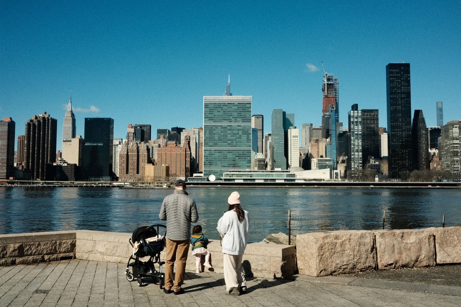 Família com carrinho de bebê e criança pequena aproveitando a vista do skyline de Nova York em um parque à beira-mar num dia ensolarado.