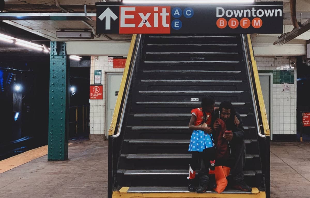 Vater und Kind in einer U-Bahn-Station in New York City, gemeinsam am Fuß einer Treppe unter auffälligen U-Bahn-Schildern sitzend.