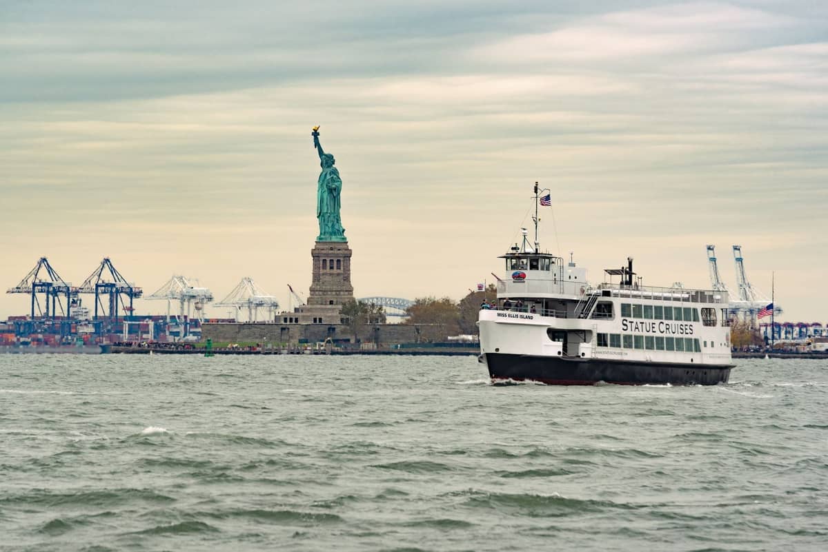 Eine Fähre von Statue Cruises fährt auf die Statue of Liberty zu, die Statue und der Hafen sind unter einem bewölkten Himmel sichtbar.