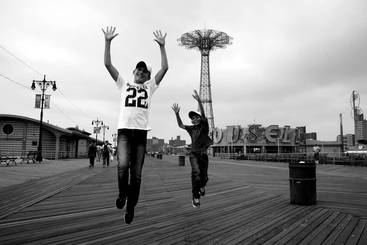 Zwei Kinder springen auf der Strandpromenade von Coney Island mit dem Parachute Jump und einem Karussellschild im Hintergrund.