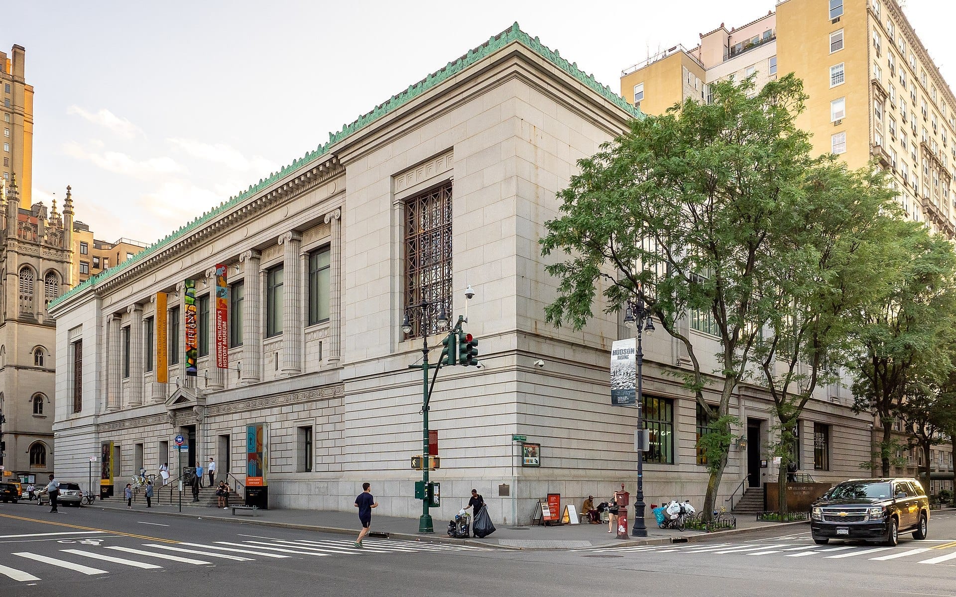 Vista angolare della facciata neoclassica in pietra della New-York Historical Society, con marciapiedi alberati, striscioni del museo e persone che camminano in una giornata di sole.