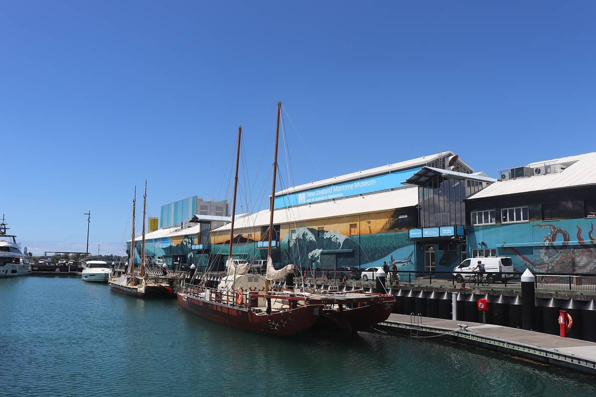 Außenansicht des New Zealand Maritime Museum in Auckland mit buntem Wandgemälde an der Fassade, Museumsschild und historischen Segelschiffen am Kai.