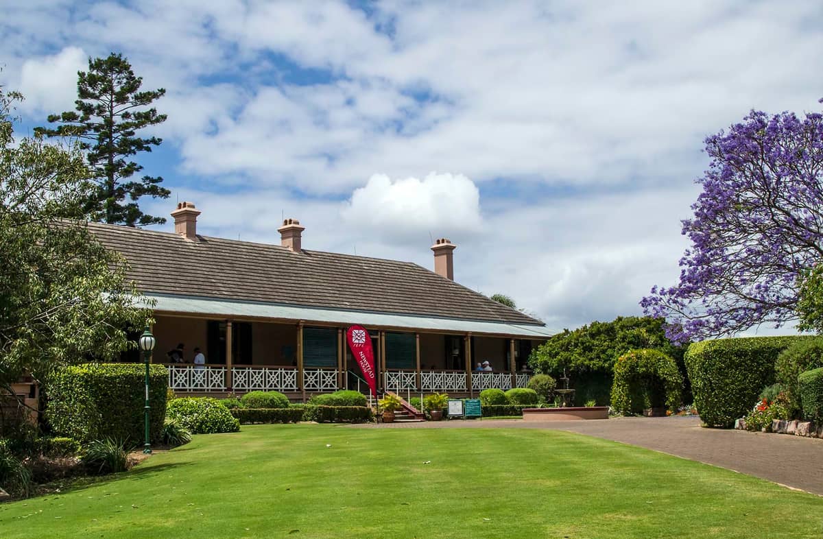 Newstead House, Brisbanes ältestes erhaltenes Wohnhaus, von vorne mit breiter Veranda, kolonialen Geländern, Rasenfläche und umliegendem Garten unter einem teils bewölkten Himmel.