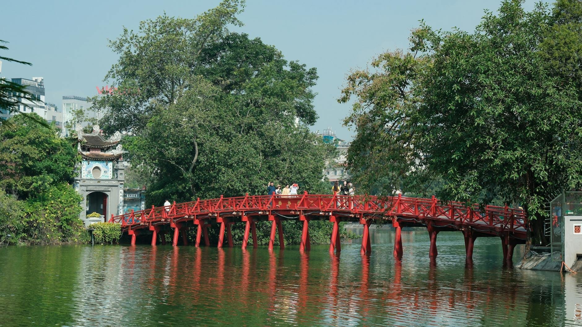 Panoramic view of the iconic red The Huc Bridge leading to Ngoc Son Temple on a lush green island in Hanoi’s Hoan Kiem Lake, with visitors crossing.