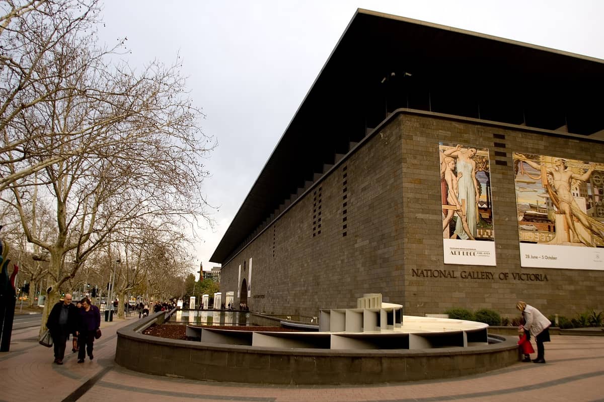 Vista oblicua del edificio NGV International sobre St Kilda Road, con su arquitectura modernista, fachada de piedra, entrada principal y espejo de agua. Personas caminan por los senderos arbolados del exterior.