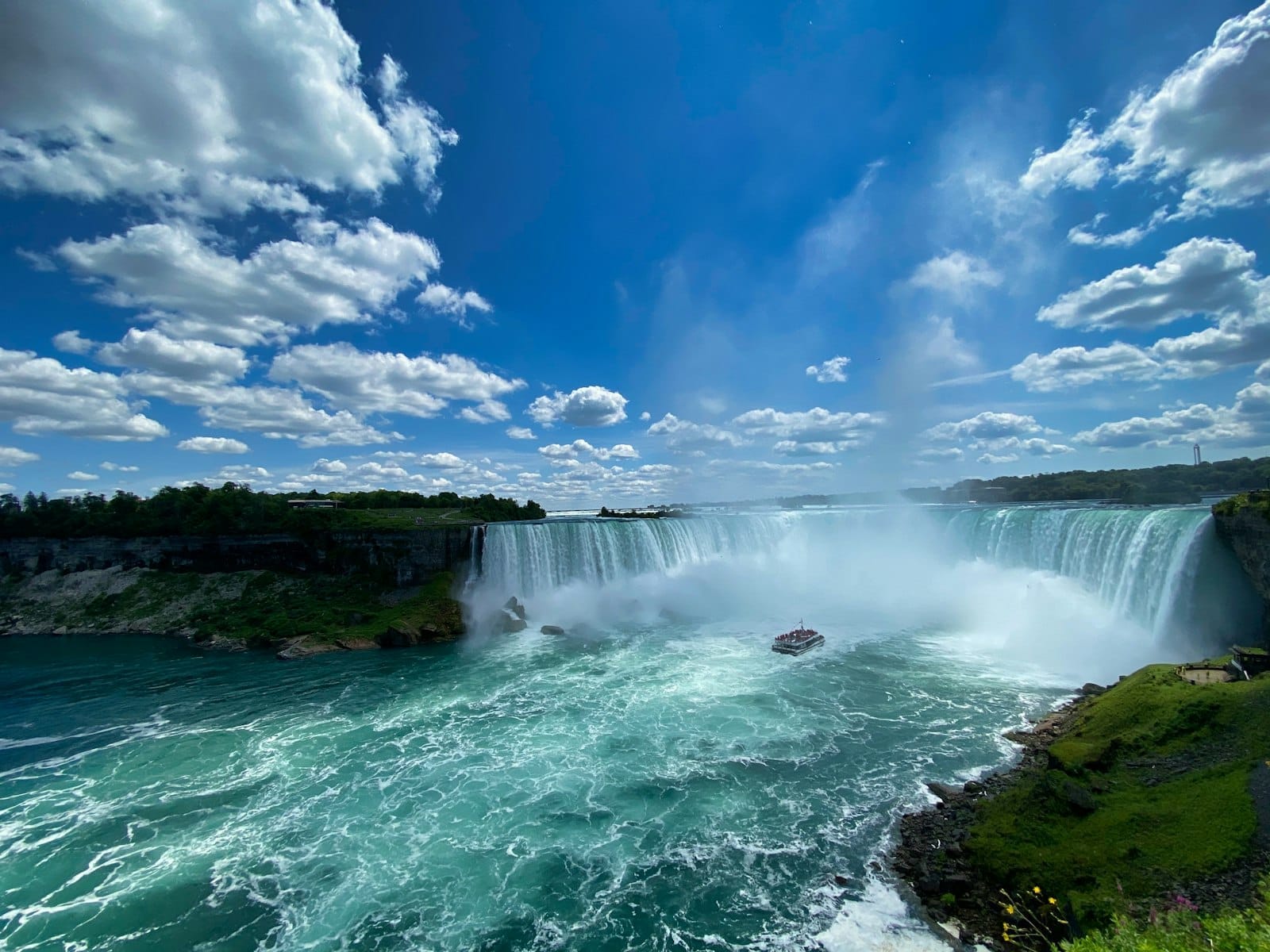 วิวกว้างของน้ำตก Niagara พร้อมน้ำสีเทอร์ควอยซ์ ละอองน้ำที่ลอยขึ้น เรือนำเที่ยวใกล้น้ำตก ภูมิทัศน์สีเขียว และท้องฟ้าสีฟ้าเต็มไปด้วยเมฆ