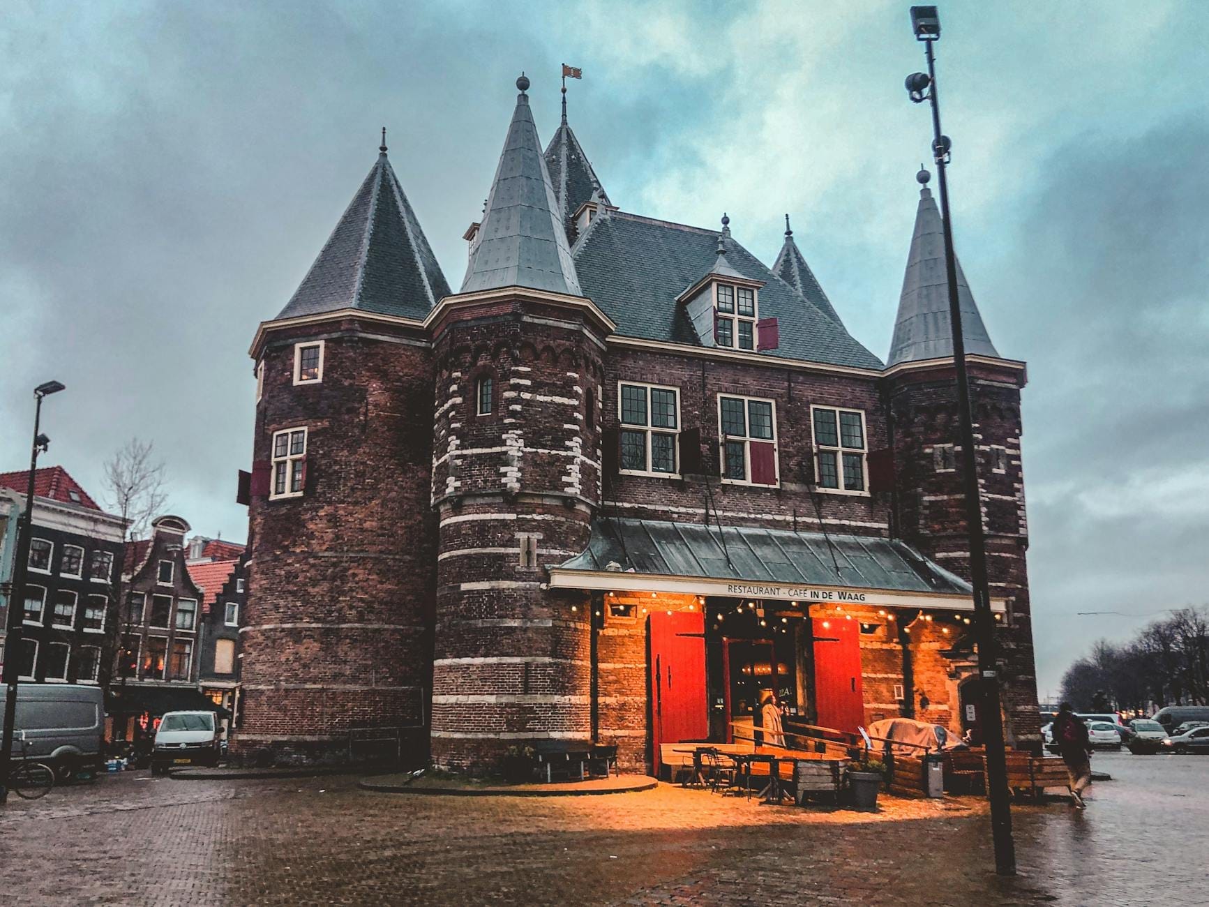 Vista del histórico edificio De Waag en la plaza Nieuwmarkt de Ámsterdam al atardecer, con las luces encendidas y el adoquinado en primer plano.
