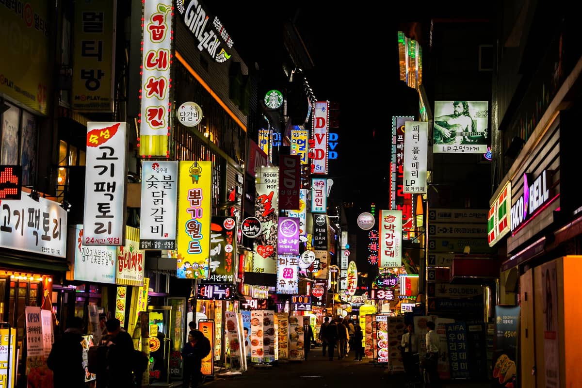 Rua movimentada do distrito noturno de Seul à noite, repleta de letreiros de néon brilhantes em coreano e pessoas caminhando em frente a bares e restaurantes.