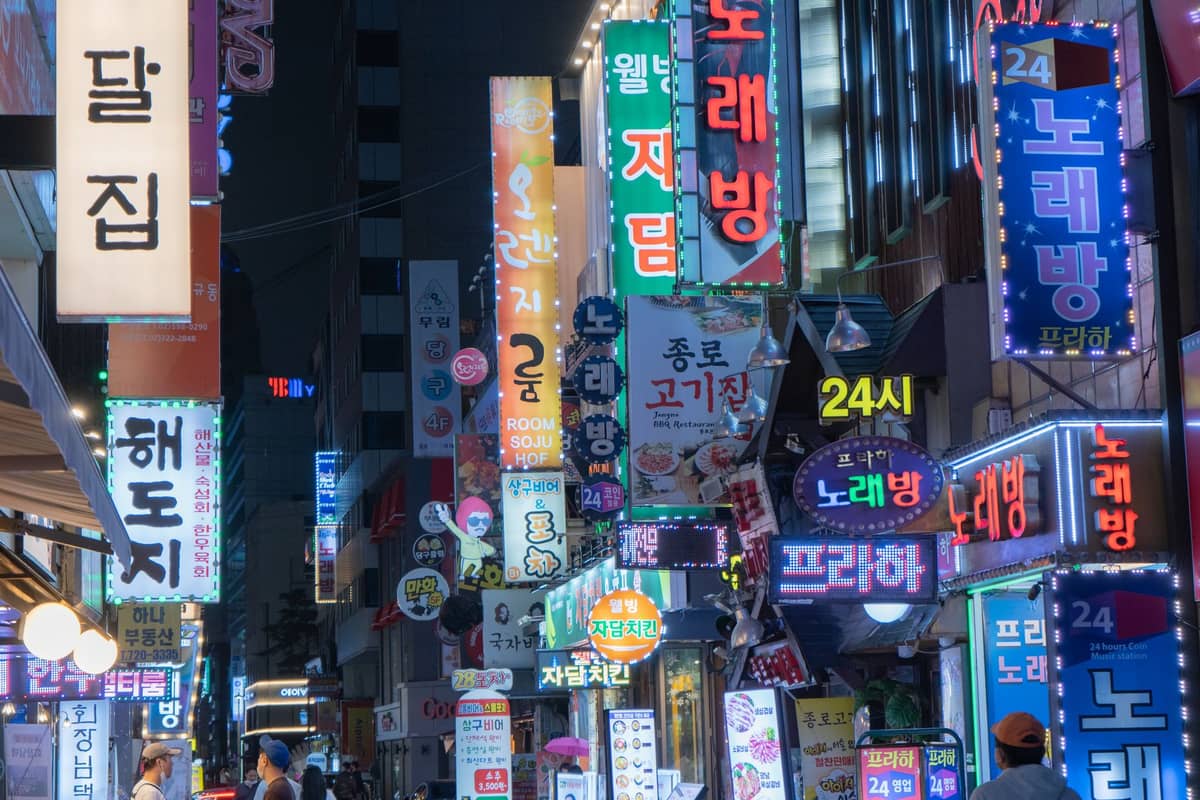 Cena noturna movimentada em Seul com multidões caminhando sob letreiros de neon de bares e karaokês em coreano, capturando a atmosfera vibrante dos bairros noturnos mais populares da cidade.