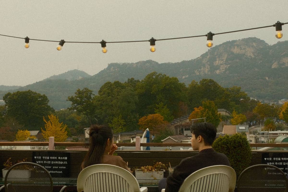 Um casal sentado ao ar livre em um bar no terraço em Seul com luzes de cordão acima e folhagem outonal ao fundo.
