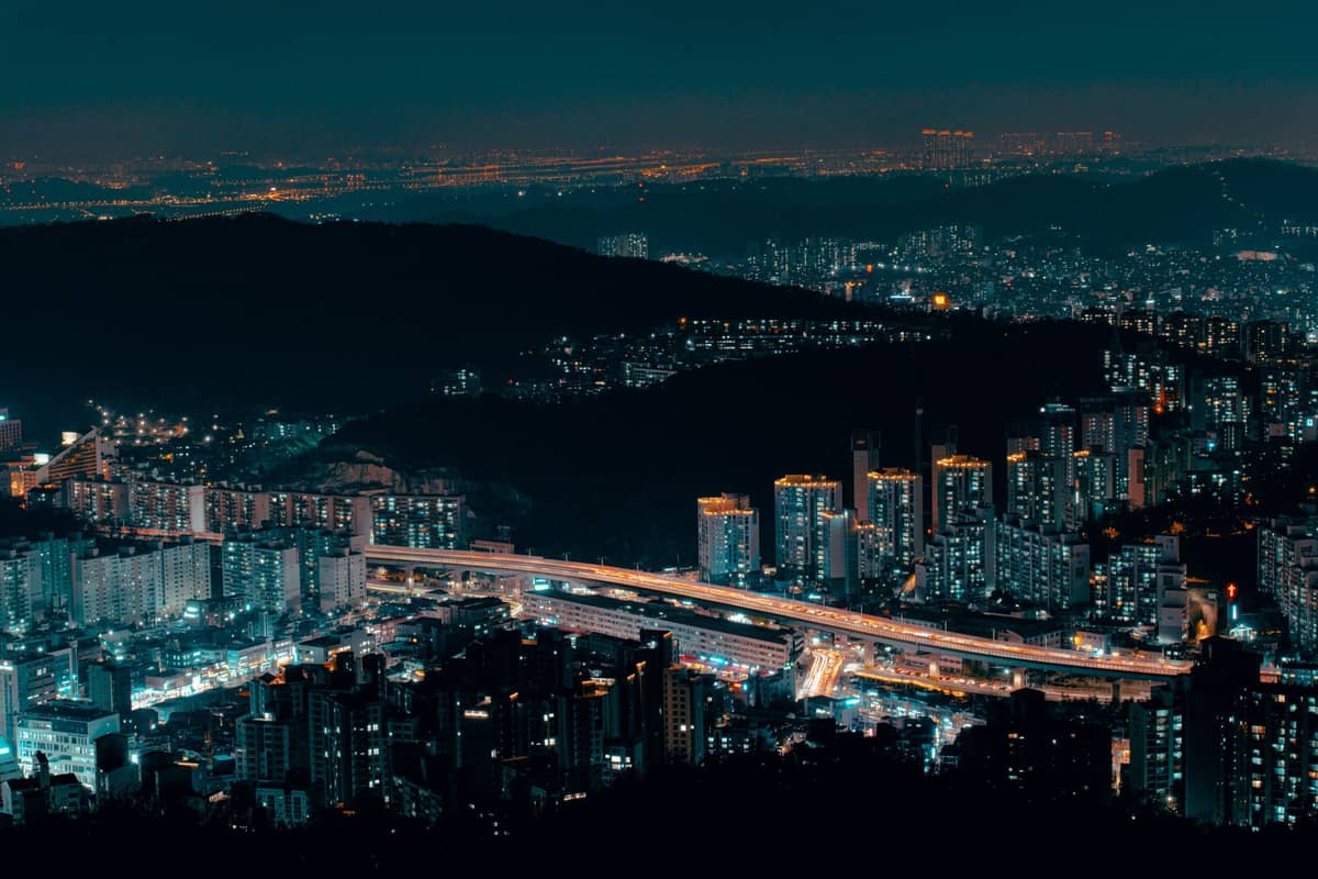 Vista panorâmica noturna de Seul com as luzes da cidade iluminando arranha-céus, ruas movimentadas e colinas ao redor sob um céu azul escuro.