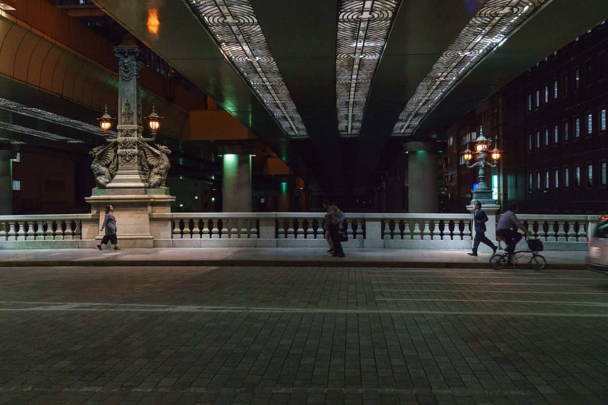 Street-level view of Nihonbashi Bridge at night, showing its iconic dragon statues, historic stone railings, and people walking beneath a highway overpass.