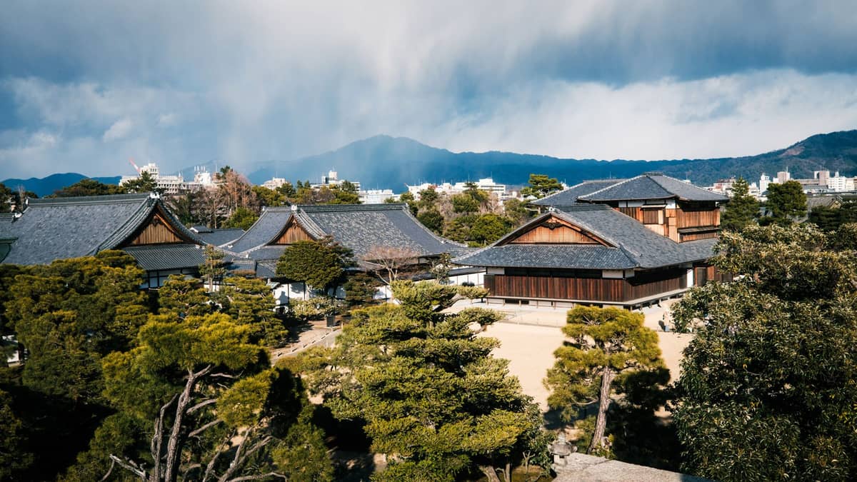Nijō Castle Palace in Kyoto, Japan, seen from above with traditional rooftops, gardens, and the cityscape with mountains in the background on a cloudy day.