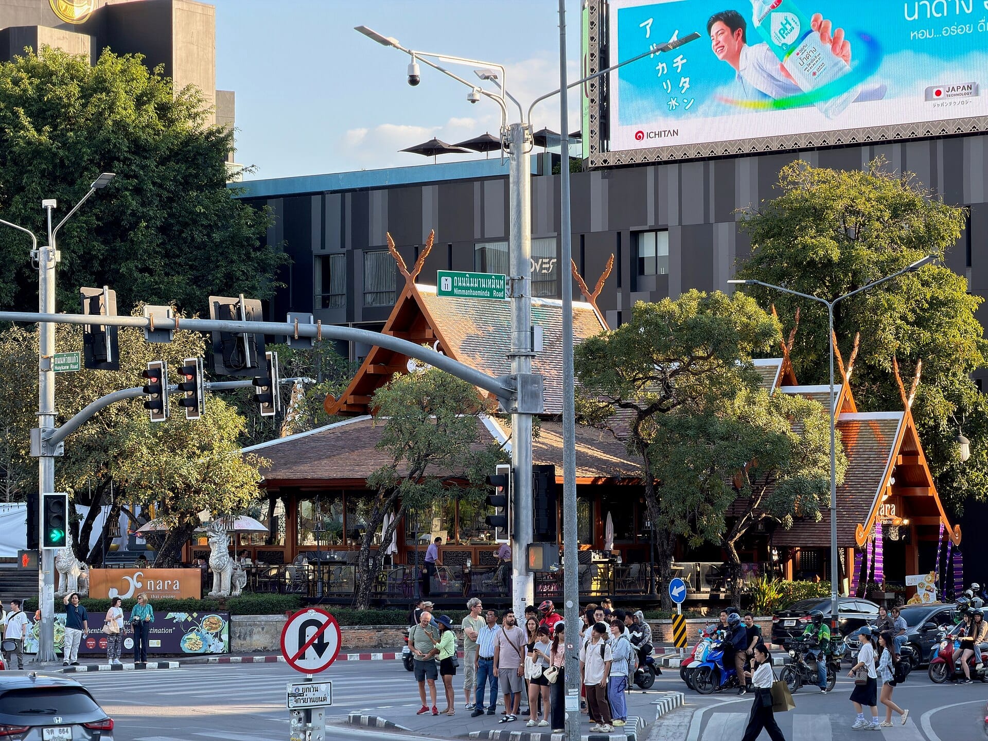Concurrida intersección en la calle Nimmanhaemin en Chiang Mai con edificios modernos, arquitectura tradicional tailandesa, semáforos y grupos de personas cruzando la calle.