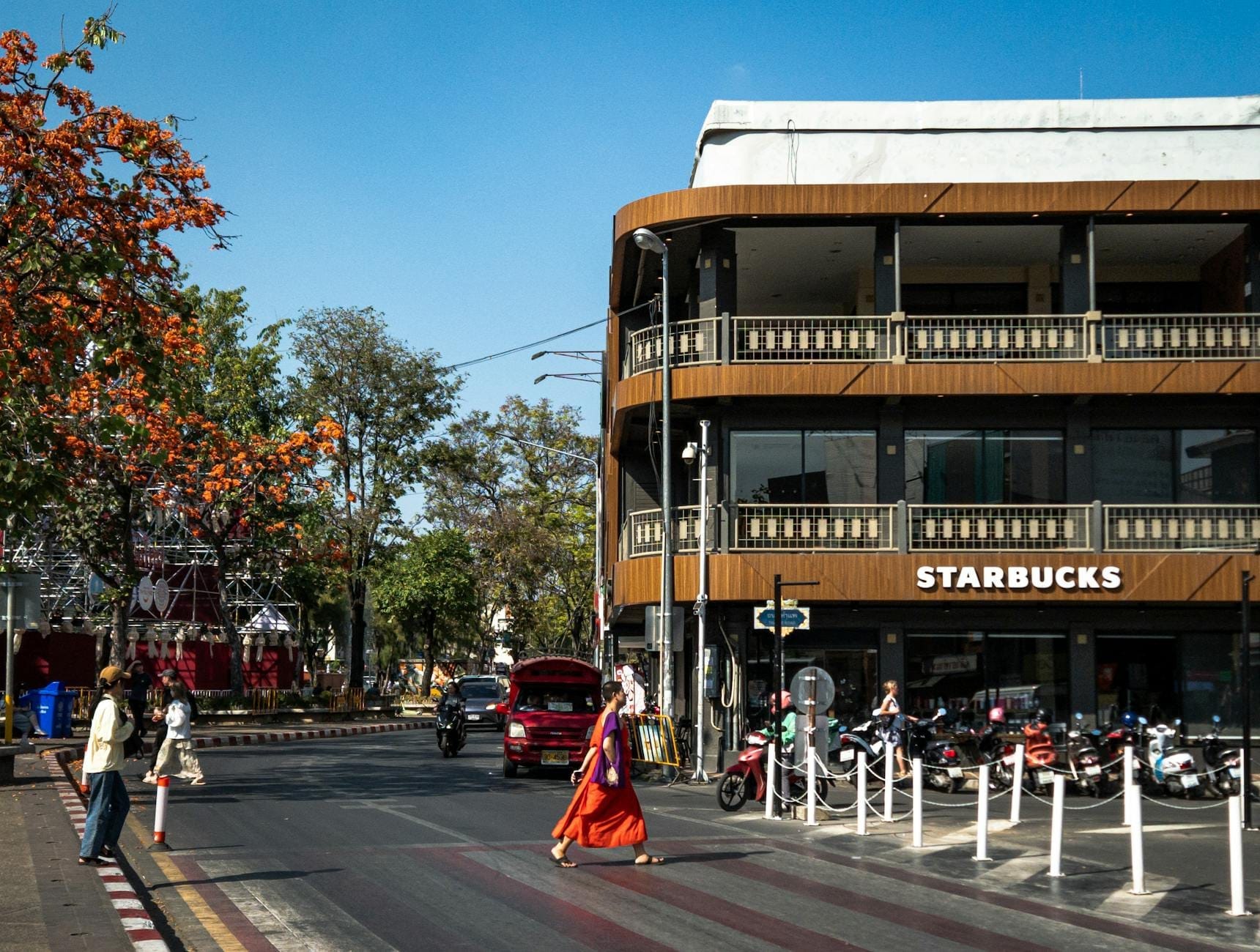 Vista de la calle Nimmanhaemin con un moderno edificio de Starbucks, motocicletas estacionadas afuera, peatones cruzando y árboles con flores naranjas bajo un cielo azul.