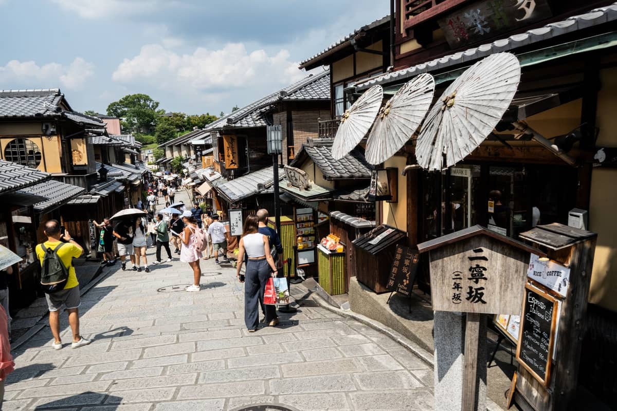 Daytime view down Ninenzaka slope in Kyoto, showing stone steps, traditional wooden buildings, teahouses, shopfronts, and parasols with people walking along the historic street.