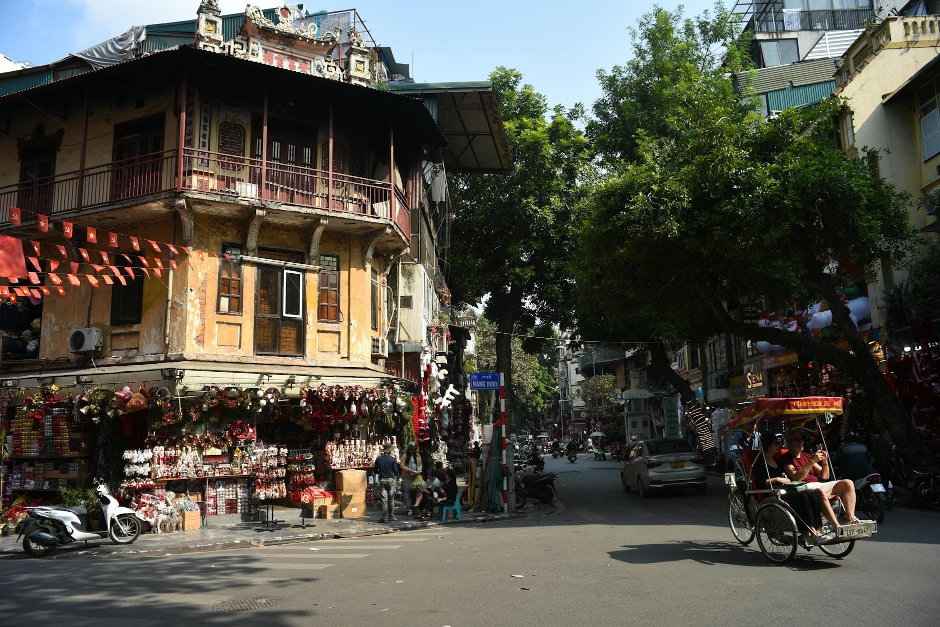 A bustling Hanoi street corner with colonial buildings, street vendors, motorcycles, and a cycle rickshaw in daylight.