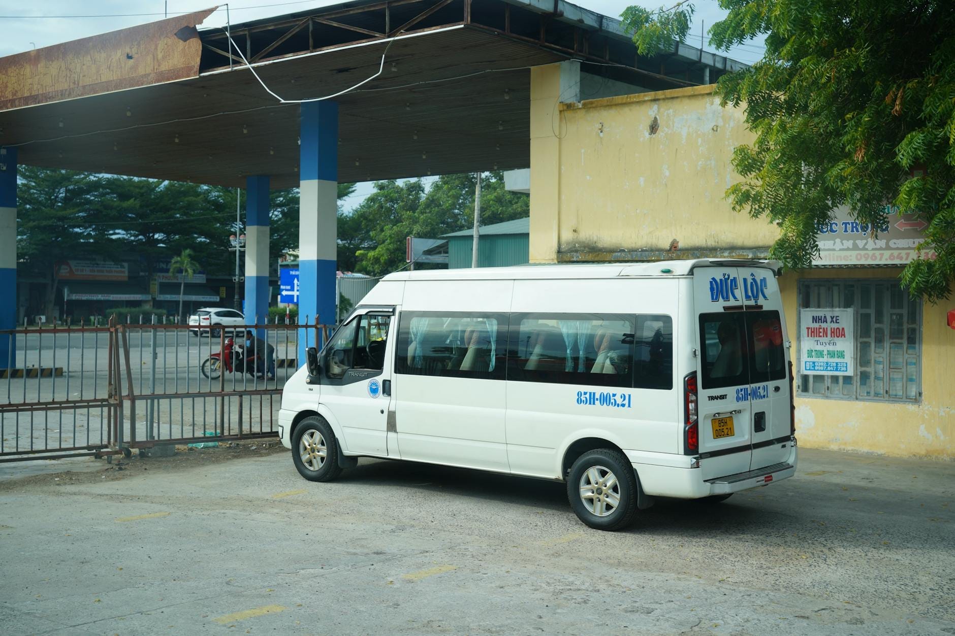 A white passenger van parked at a modest bus station with a yellow wall and some signage, typical for regional travel in Vietnam.