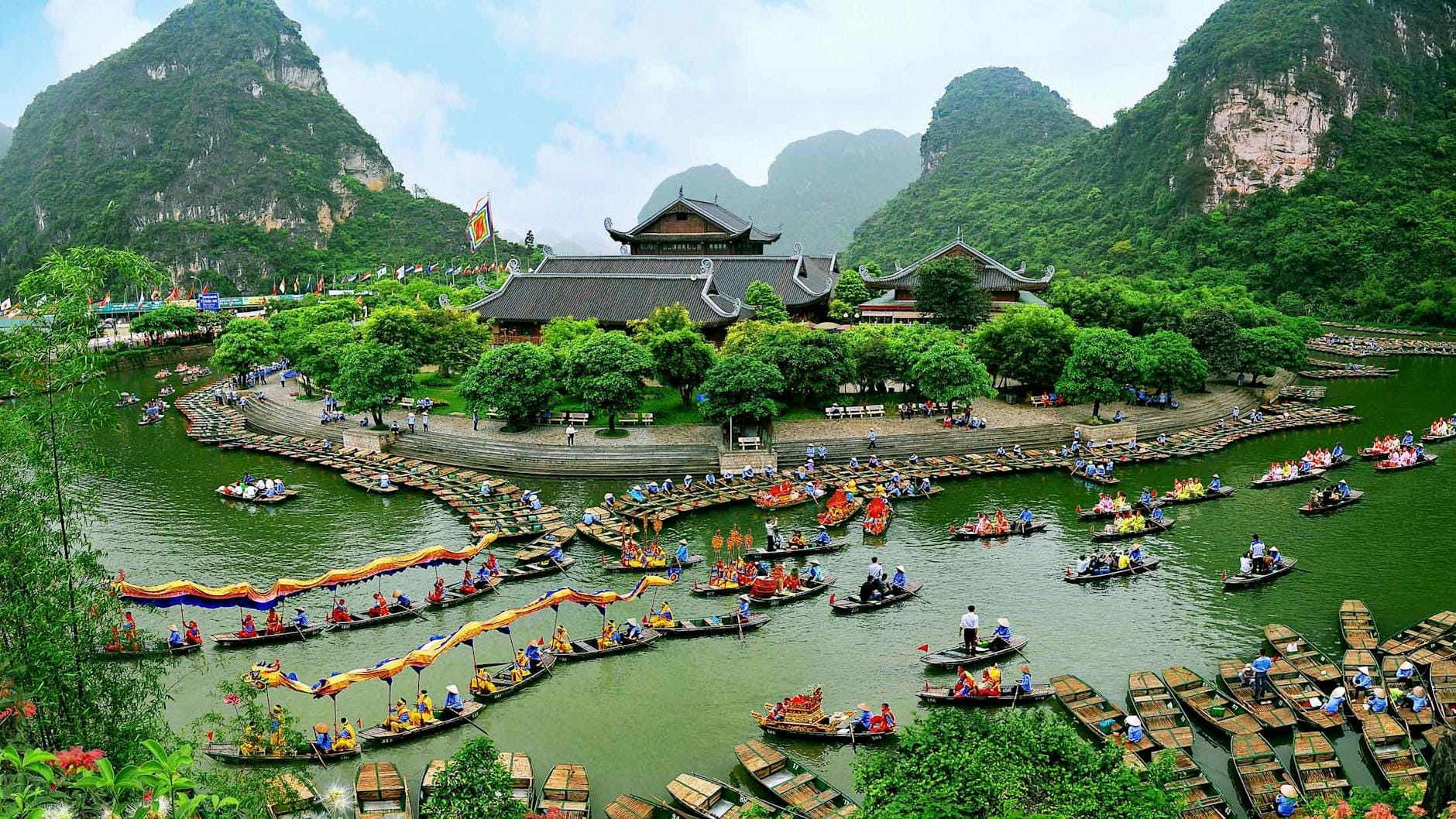 A wide view of Tràng An with dozens of rowboats on a green river, a large traditional building, lush mountains, and caves in the background.