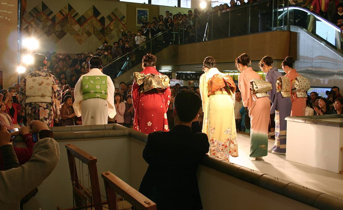A kimono fashion show inside the Nishijin Textile Center, with models in colorful kimono on stage and a large audience watching.