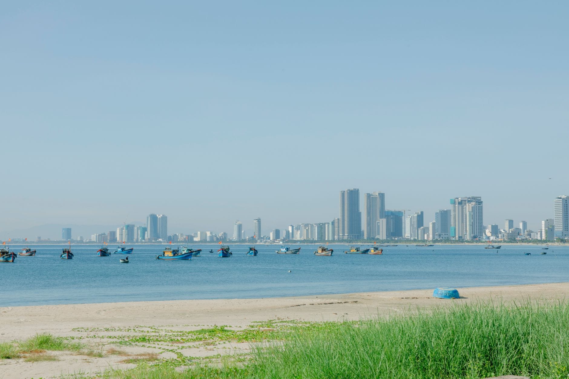 ชายหาดทรายกว้าง มีหญ้าขึ้นเป็นหย่อมๆ ด้านหน้า น้ำทะเลสีฟ้า เรือประมงเล็กๆ และวิวเมืองดานัง ตึกสูงไกลๆ ใต้ท้องฟ้าแจ่มใส